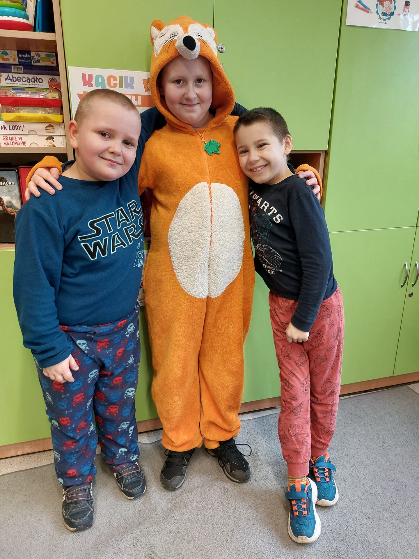 3 małych chłopców stoi w piżamach, chłopiec w środku ma piżamę -kombinezon w kolorze pomaranczowym (lis)