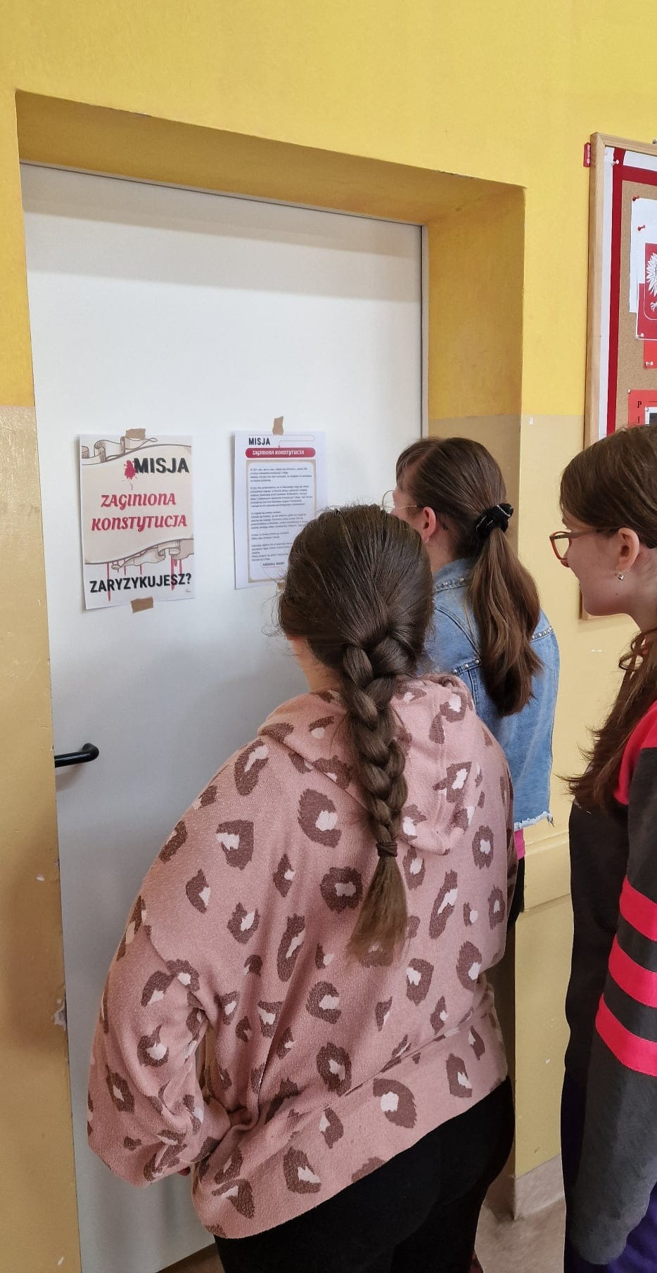 3 uczennice stoją przed drzwiami i czytają naklejoną na nie kartkę z zadaniem