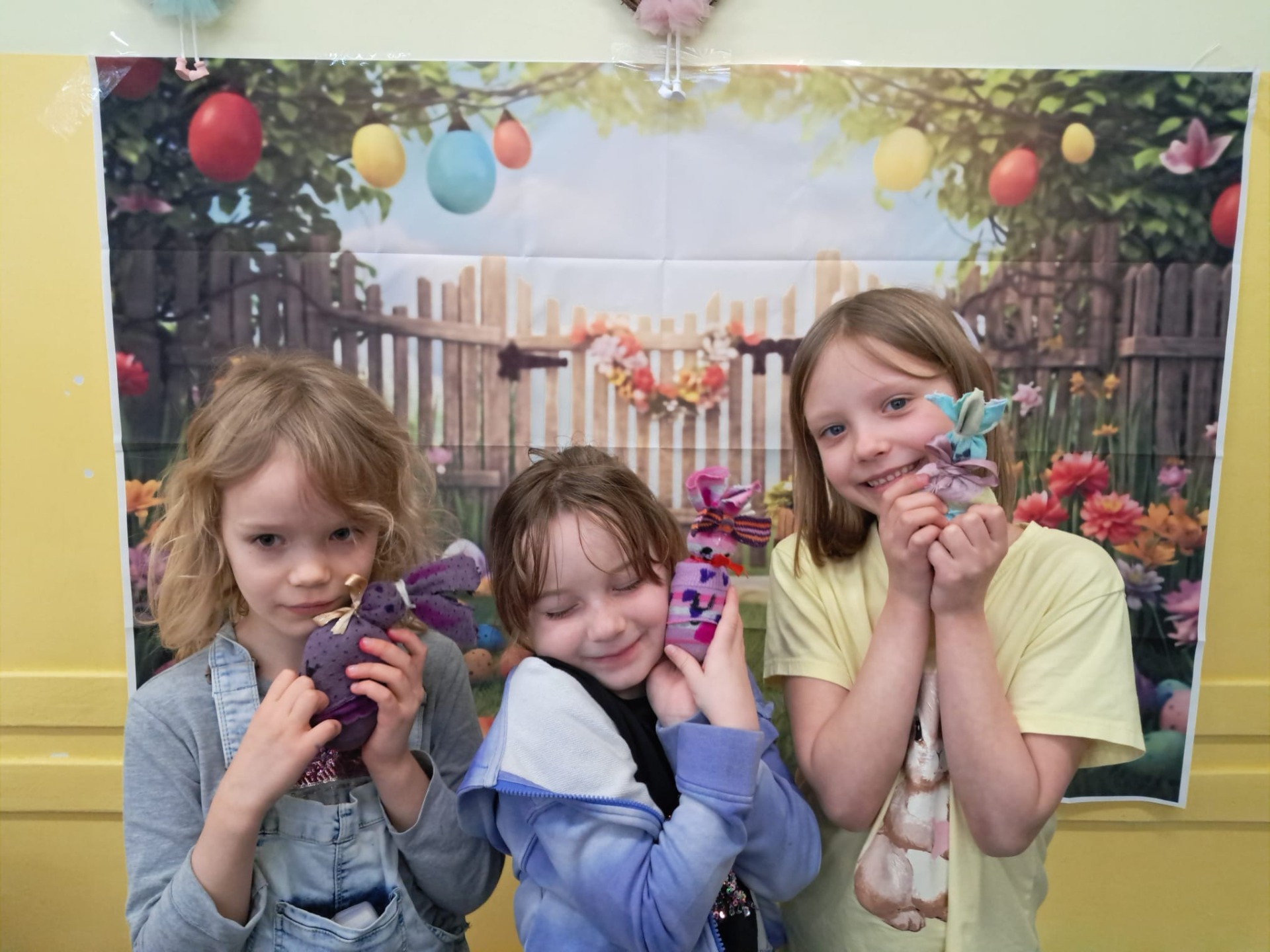 Trzy uczennice pozują do zdjęcia na tle ozdób wielkanocnych takich jak kwiaty i kolorowe pisanki. Dziewczynki trzymają w rękach wielkanocne zające.