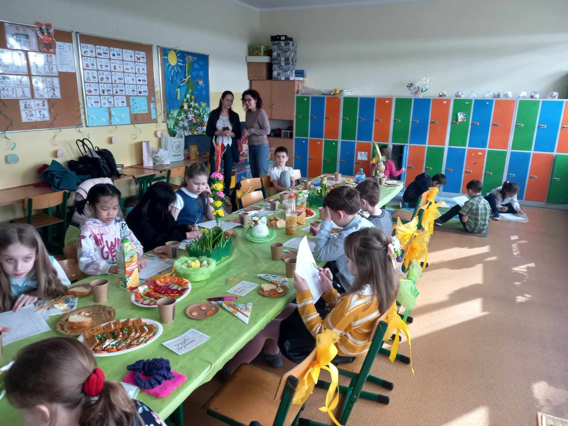 Uczniowie w klasie siedzą przy złączonych ławach nakrytych zielonym obrusem. Na tak urządzonym wielkanocnym stole stoi poczęstunek na talerzach, dzieci siedzą i zajadają się smakołykami. W głębi klasy widać szafki uczniów w kolorze niebieskim, pomarańczowym i zielonym obok których stoją dwie wychowawczynie.