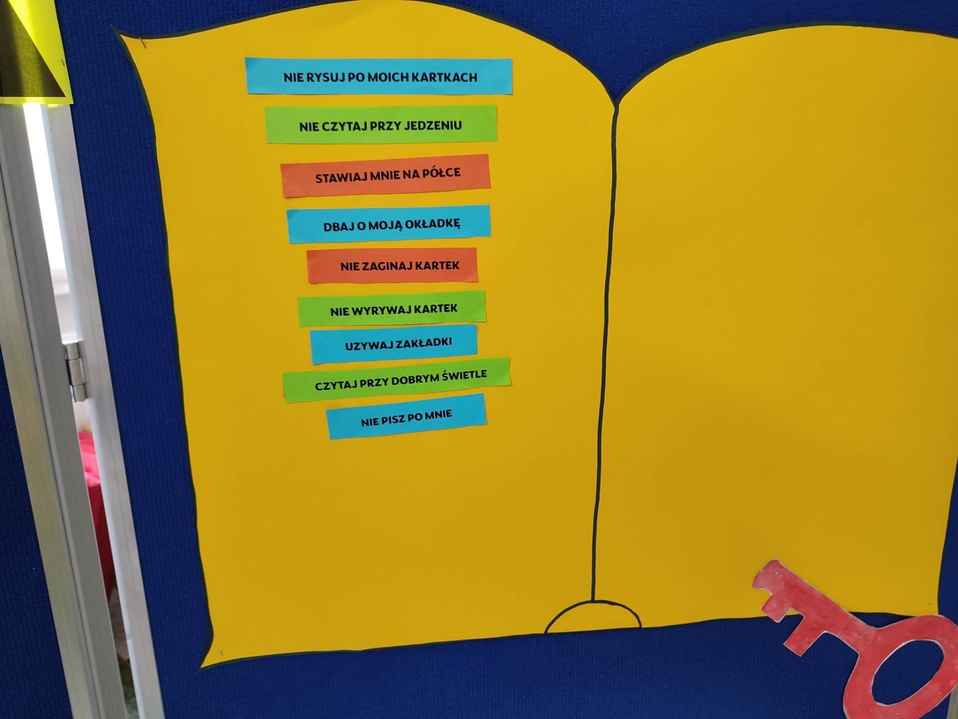 Na niebieskim tle przymocowano żółtą planszę w kształcie książki z zasadami wypożyczania książek.