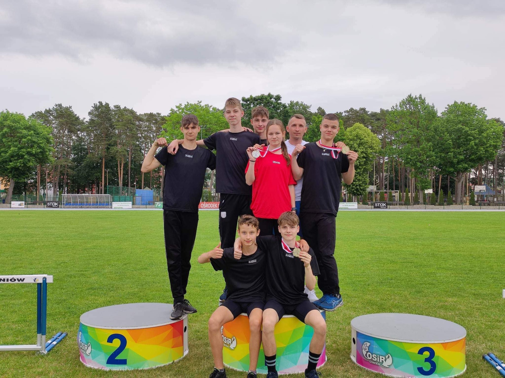Siedmioro uczniów z medalami  i trener na podium podczas Mistrzostw Województwa w Lekkiej Atletyce klas 7-8.