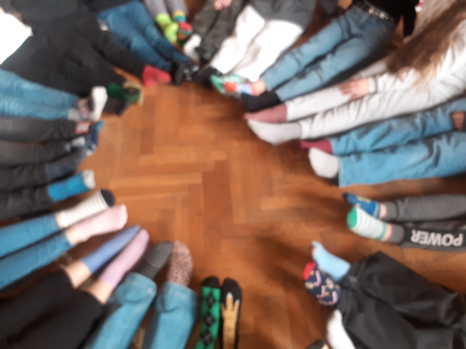 Fotografia przedstawia stopy uczniów naszej szkoły, którzy założyli dziś kolorowe skarpety nie do pary. Uczniowie siedzą w kole, na sali gimnastycznej.