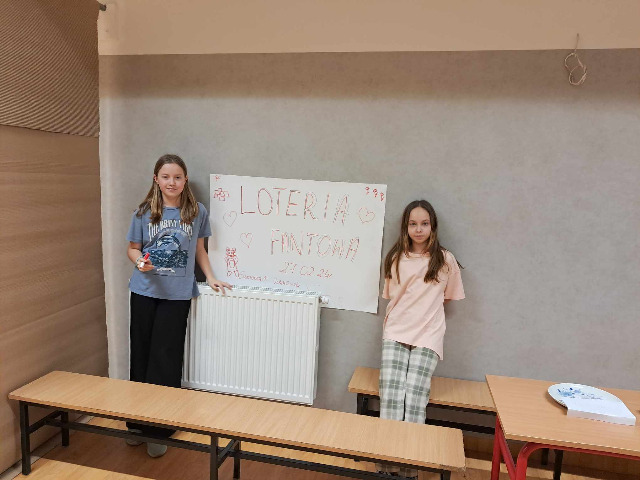 Dwie dziewczynki ubrane w jasne kolory stoją na tle szarej ściany. Pomiędzy nimi powieszony jest plakat z napisem loteria fantowa. Przed nimi stoi ławka szkolna.