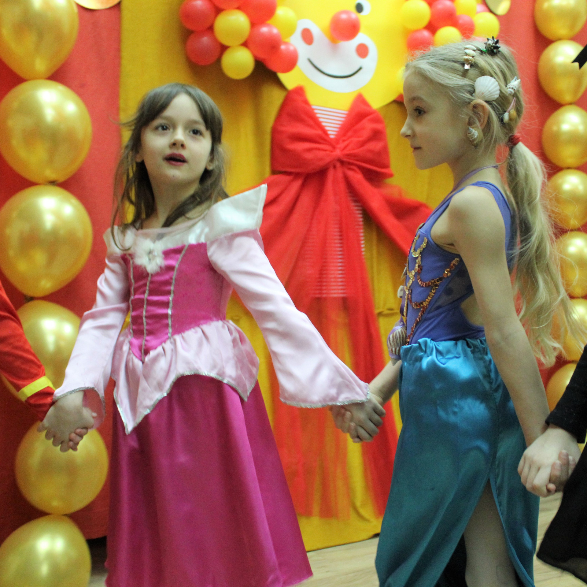 Dwie dziewczynki w strojach księżniczek na balu karnawałowym, tańczą i śpiewają trzymając się za ręce.