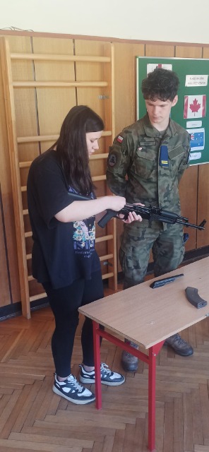 Uczennica trzyma w ręku broń palną, obok niej stoi umundurowany chłopiec. Na stole, który stoi na przeciwko nich leżą elementy broni.