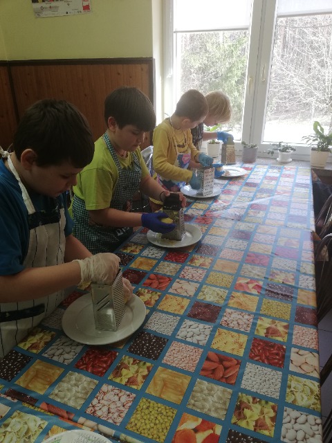 Stołówka szkolna. 4 uczniów, stoją przy stole, na tarkach tarkują ziemniaki .

