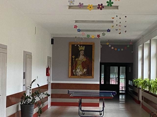 Pierwsze zdjęcie przedstawia korytarz szkoły. Na środku korytarza stoi stół ping-pongowy, od lamp zwisają dekoracje, kwiaty wiosenne i motyle. Na wprost na ścianie wisi portret Świętej Królowej Jadwigi patronki SP w Dydni. Na parapetach ustawione są kwiaty.