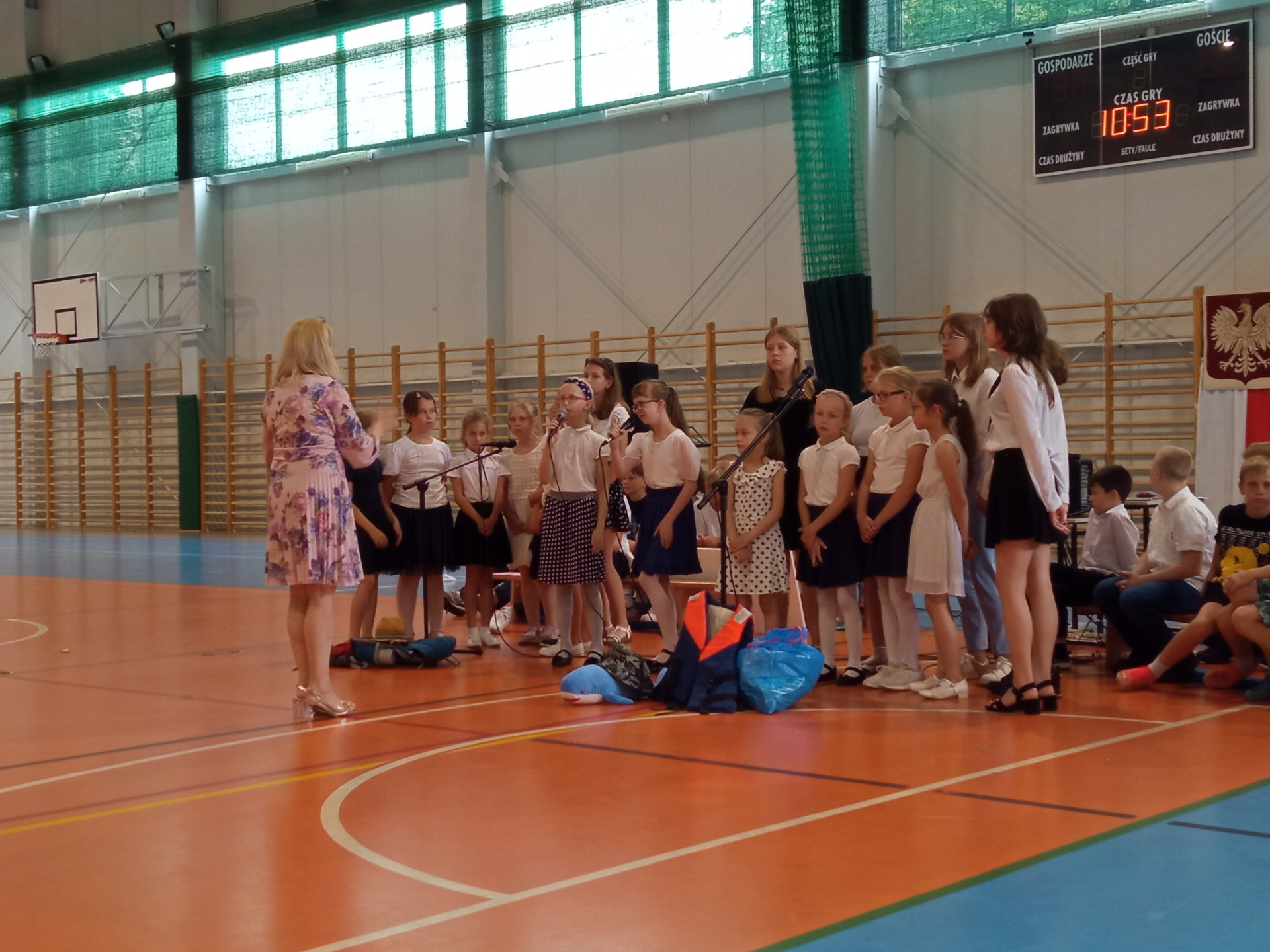 Grupa uczniów śpiewa utwory muzyczne na hali sportowej pod batutą nauczyciela muzyki.