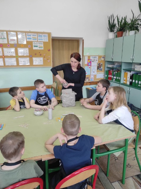 Dorosła kobieta pokazuje 6 dzieci siedzącym wokół stołu sposób lepienia  kubka w glinie