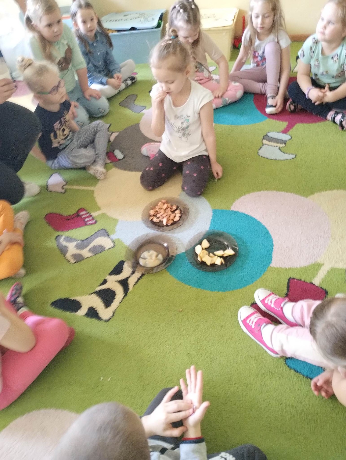 Dzieci siedzą w kole na kolorowym dywanie w sali przedszkolnej i biorą udział w warsztatach w ramach projektu edukacyjnego "Gramy zmysłami"