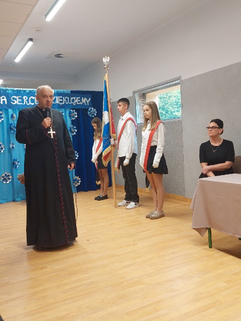 Po lewej stronie stoi duchowny, w ręku trzyma mikrofon. Po jego lewej stronie stoi 3 uczniów ubranych na biało-czarno, na ramieniu mają biało-czerwone szarfy. Przy stoliku siedzi kobieta.
