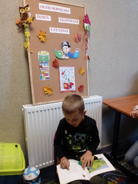 Chłopiec siedzi i na kolanach trzyma otwartą książkę. Za nim tablica korkowa    z napisem Ogólnopolski Dzień Głośnego Czytania.