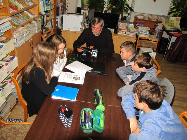Przy stole siedzi nauczyciel, po jego prawej 2 uczennice, po lewej 3 uczniowie. Na stole leżą piórniki i książki. Uczniowie pochylają się nad książką.