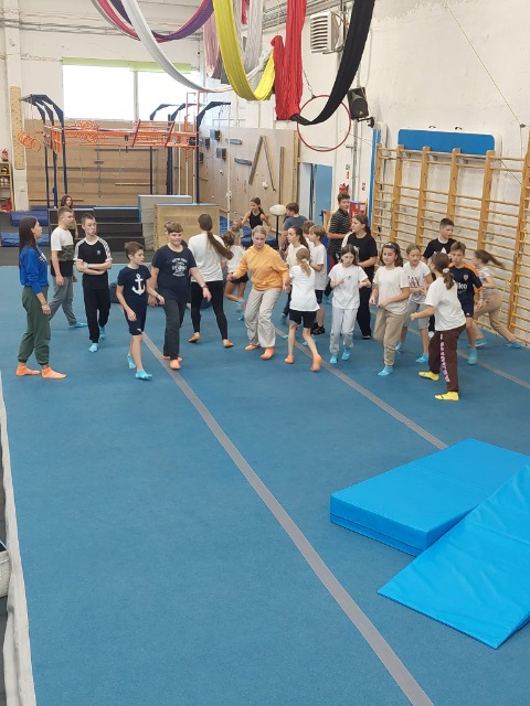 Grupa uczniów stoi na niebieskim materacu w dużej sali gimnastycznej.