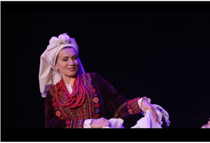 Zdjęcie aktorki w trakcie spektaklu, na głowie biała chusta, czerwone korale i zdobiona bordowa marynarka. 